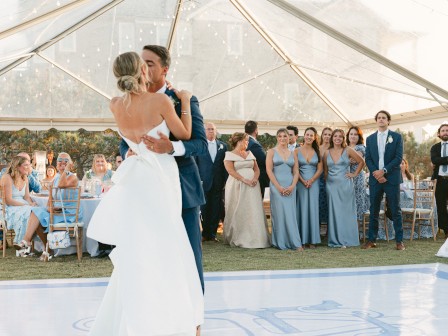 A couple dances at an outdoor wedding reception under a tent, with guests watching and wearing formal attire.