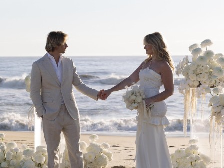 A couple in wedding attire stands on a beach, holding hands. The decor includes white flowers, and the ocean is in the background.