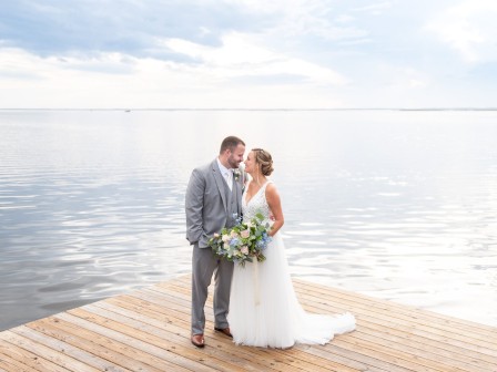 A couple in wedding attire stands on a wooden dock by a calm lake, with a serene sky reflecting in the water.