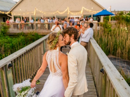 A bride and groom share a kiss on a wooden walkway with guests in the background at an outdoor wedding venue.