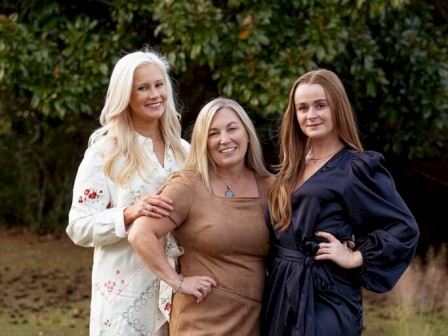 Three women are posing outdoors, smiling, with trees in the background.