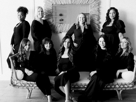 A black-and-white photo of nine women in black dresses, posing together on a couch and rug, with a mirror on the wall behind them.