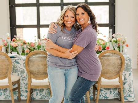 Two people are hugging and smiling in front of a decorated table with flowers and chairs.