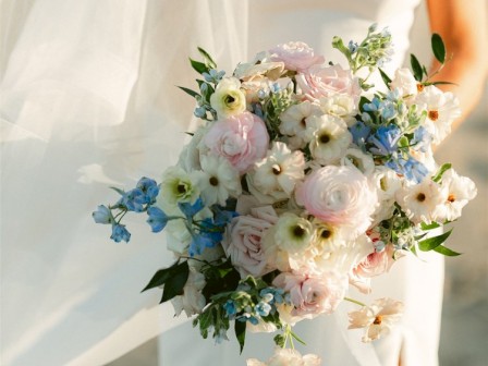 A bride in a white dress holding a bouquet of pastel flowers, featuring pink, white, and blue blooms, with a delicate veil.