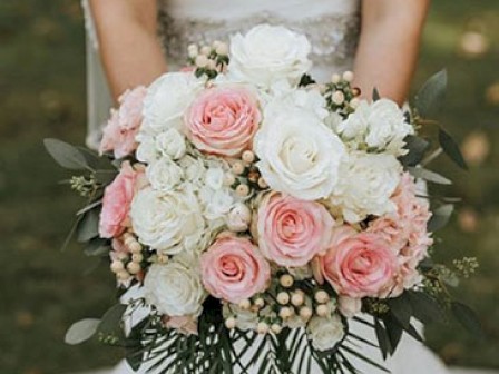 A bride holds a bouquet of pink and white roses with green foliage, dressed in a white gown, showcasing an elegant wedding arrangement.