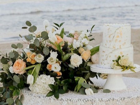 A beachside setting with a floral arrangement and a two-tiered white cake on a decorated table, capturing a serene, elegant atmosphere.