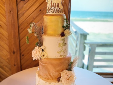 A multi-tiered wedding cake with floral decorations and a silhouette topper, set in front of a beach view background.