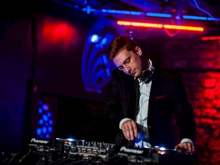 A DJ in a dark club setting, wearing headphones, adjusts the mixer. Neon lights illuminate the scene with a vibrant ambiance.