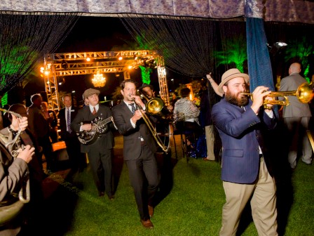 A band is playing at an outdoor event with musicians walking and performing under decorative lighting and a canopy.