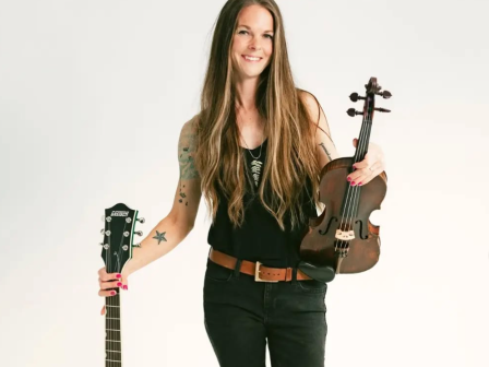 A person holding a guitar and a violin, wearing a black outfit, standing against a plain background.