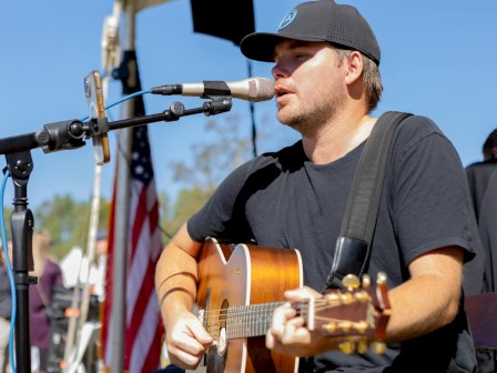 A person in a cap sings and plays an acoustic guitar on stage, with a microphone and a U.S. flag visible in the background.