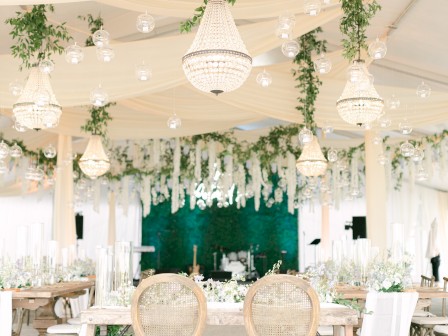 An elegantly decorated venue features draped fabric, chandeliers, string lights, and abundant greenery, creating a luxurious and inviting atmosphere.