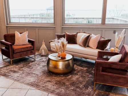 The image shows a cozy living room with a sofa, two armchairs, a round metallic coffee table, pillows, and decorative items on a rug.