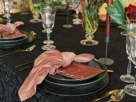 A beautifully set dining table with black tablecloth, elegant candles, pink napkins, decorative flowers, and glassware.