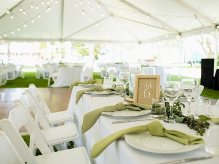 A beautifully set table under a tent with white chairs, green napkins, glasses, and a 