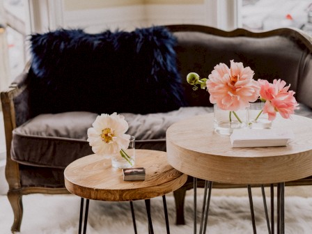 A cozy seating area with a gray couch, dark blue pillows, and round wooden side tables decorated with pink and white flowers in vases.