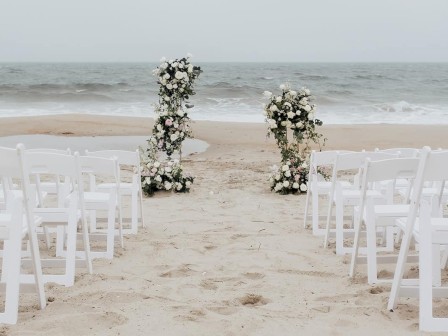 A beach wedding setup with white chairs and floral arrangements facing the ocean.