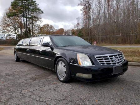 A black stretch limousine is parked on a paved area with trees and a cloudy sky in the background.